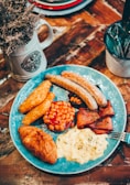 A hearty breakfast plate features sausages, hash browns, baked beans, bacon, scrambled eggs, and a croissant, artfully arranged on a blue decorative plate. Nearby, a rustic mug filled with dried flowers and a cup holding cutlery sit on a wooden table, contributing to a cozy, homely atmosphere.