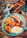 A hearty breakfast plate features sausages, hash browns, baked beans, bacon, scrambled eggs, and a croissant, artfully arranged on a blue decorative plate. Nearby, a rustic mug filled with dried flowers and a cup holding cutlery sit on a wooden table, contributing to a cozy, homely atmosphere.