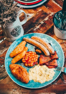 A hearty breakfast plate features sausages, hash browns, baked beans, bacon, scrambled eggs, and a croissant, artfully arranged on a blue decorative plate. Nearby, a rustic mug filled with dried flowers and a cup holding cutlery sit on a wooden table, contributing to a cozy, homely atmosphere.
