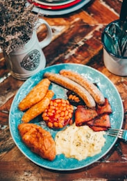 A hearty breakfast plate features sausages, hash browns, baked beans, bacon, scrambled eggs, and a croissant, artfully arranged on a blue decorative plate. Nearby, a rustic mug filled with dried flowers and a cup holding cutlery sit on a wooden table, contributing to a cozy, homely atmosphere.