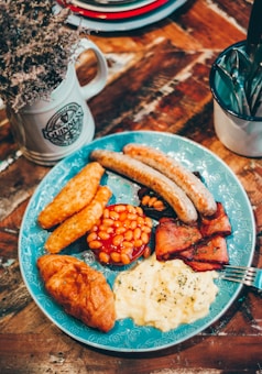 A hearty breakfast plate features sausages, hash browns, baked beans, bacon, scrambled eggs, and a croissant, artfully arranged on a blue decorative plate. Nearby, a rustic mug filled with dried flowers and a cup holding cutlery sit on a wooden table, contributing to a cozy, homely atmosphere.