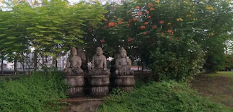Three stone statues are seated on pedestals in an outdoor garden setting, surrounded by lush green foliage and vibrant flowering plants. Each statue bears traditional attire and is positioned under a canopy of trees and shrubs.