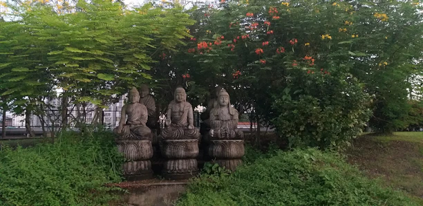 Three stone statues are seated on pedestals in an outdoor garden setting, surrounded by lush green foliage and vibrant flowering plants. Each statue bears traditional attire and is positioned under a canopy of trees and shrubs.
