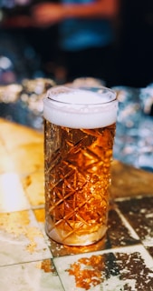 A glass of amber-colored beer with a frothy head sits on a reflective, textured table. The glass has a unique geometric pattern, adding elegance to the scene. The background is blurred, with warm lighting suggesting a cozy atmosphere.
