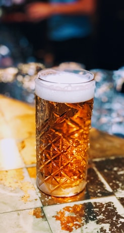 A glass of amber-colored beer with a frothy head sits on a reflective, textured table. The glass has a unique geometric pattern, adding elegance to the scene. The background is blurred, with warm lighting suggesting a cozy atmosphere.