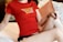 A person wearing a red t-shirt with the text 'Real Poker' printed in yellow, sitting on a bed with white sheets and brown pillows. The person's pose is casual and relaxed, with one leg bent and an arm resting on the knee.