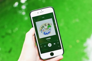 A hand is holding a smartphone displaying a music app with album art and music controls visible on the screen. The background consists of a vibrant green surface.