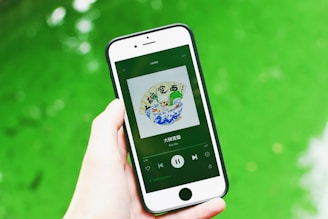 A hand is holding a smartphone displaying a music app with album art and music controls visible on the screen. The background consists of a vibrant green surface.