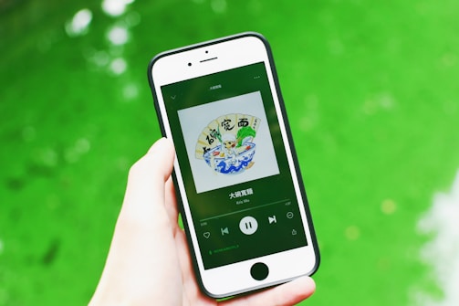 A hand is holding a smartphone displaying a music app with album art and music controls visible on the screen. The background consists of a vibrant green surface.