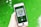 A hand is holding a smartphone displaying a music app with album art and music controls visible on the screen. The background consists of a vibrant green surface.