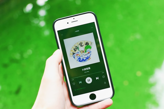 A hand is holding a smartphone displaying a music app with album art and music controls visible on the screen. The background consists of a vibrant green surface.
