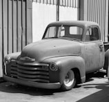 A vintage pickup truck with a classic design, featuring a rounded hood and prominent front grille. The truck has a matte finish and appears to be parked next to a corrugated metal wall in an industrial setting. The photo is in black and white, emphasizing the textures and shadows on the vehicle.