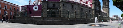 A historic city street features a large, ornate building with pink and white detailing, arched windows, and a prominent bell tower. The building is enclosed by a metal fence. Nearby, people walk and ride bicycles on the spacious stone-paved square. A small fountain with water jets adds activity to the scene. On the left, a reddish brick building with multiple windows completes the urban landscape.