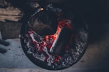 Glowing embers of charcoal and wood in a circular fire pit, surrounded by ash and charred remains.