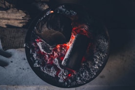 Glowing embers of charcoal and wood in a circular fire pit, surrounded by ash and charred remains.