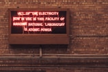 A neon sign is displayed on a brick wall, reading 'ALL OF THE ELECTRICITY NOW IN USE IN THIS FACILITY OF ARGONNE NATIONAL LABORATORY IS ATOMIC POWER'. The sign emits a reddish glow, contrasting with the earthy tones of the brick wall background.
