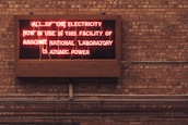 A neon sign is displayed on a brick wall, reading 'ALL OF THE ELECTRICITY NOW IN USE IN THIS FACILITY OF ARGONNE NATIONAL LABORATORY IS ATOMIC POWER'. The sign emits a reddish glow, contrasting with the earthy tones of the brick wall background.