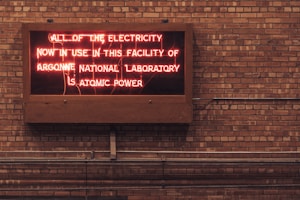 A neon sign is displayed on a brick wall, reading 'ALL OF THE ELECTRICITY NOW IN USE IN THIS FACILITY OF ARGONNE NATIONAL LABORATORY IS ATOMIC POWER'. The sign emits a reddish glow, contrasting with the earthy tones of the brick wall background.