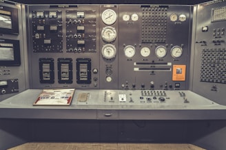 An old-fashioned control room panel filled with numerous analog dials, switches, and meters. The panel has labels such as 'Radiation', 'Reactor', and 'Interlocks', indicating its use in monitoring and controlling industrial or scientific processes. The design suggests mid-20th-century technology, with a utilitarian layout and a slight yellowed tint, giving it a vintage appearance.