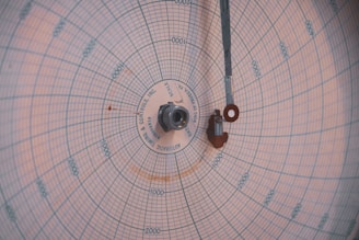 A circular chart paper with blue grid lines, marked with numerical data points and labels in a radial pattern. A central pivot with metallic components extends a thin arm across the chart. The markings indicate measurements in a systematic manner, featuring various numerical values.