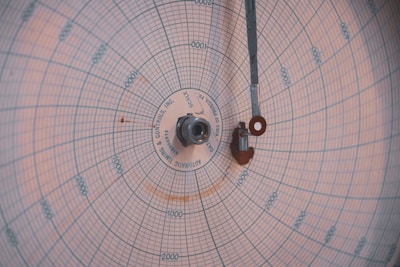 A circular chart paper with blue grid lines, marked with numerical data points and labels in a radial pattern. A central pivot with metallic components extends a thin arm across the chart. The markings indicate measurements in a systematic manner, featuring various numerical values.