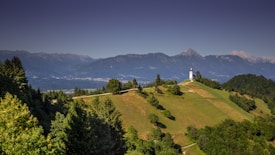 A picturesque landscape with rolling green hills, scattered trees, and a solitary white church with a steeple perched on top of a small hill. The background features vast mountainous terrain under a clear blue sky.
