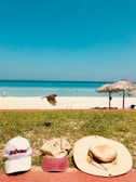 A collection of colorful trucker hats laid out on sandy beach grass with the ocean in the background.