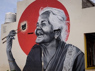 A large mural depicts an elderly woman with white hair laughing joyfully. She is holding a paintbrush, seemingly painting a bright red circle behind her. The mural is detailed in black and white, with intricate patterns on her clothing.