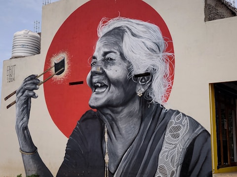 A large mural depicts an elderly woman with white hair laughing joyfully. She is holding a paintbrush, seemingly painting a bright red circle behind her. The mural is detailed in black and white, with intricate patterns on her clothing.