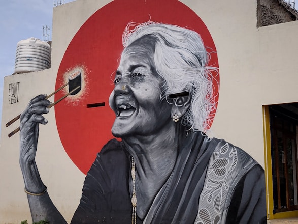 A large mural depicts an elderly woman with white hair laughing joyfully. She is holding a paintbrush, seemingly painting a bright red circle behind her. The mural is detailed in black and white, with intricate patterns on her clothing.
