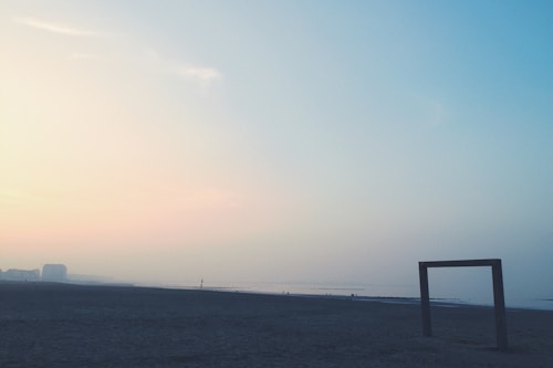 A serene beach scene with a hazy horizon during either dawn or dusk. There's a large, rectangular, framed structure positioned on the right side in the foreground. The shoreline stretches into the distance with a calm sea. The sky transitions from soft pastel hues of pink and peach on the left to light blue on the right, with a gentle gradient effect.