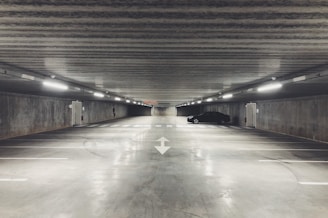 A large, empty underground parking garage with concrete walls and ceiling. There are fluorescent lights lining the ceiling, casting a dull glow throughout the space. A single black car is parked on the right side, while the rest of the parking spots are vacant. The floor is smooth and polished, with directional arrows painted on it, contributing to an overall sterile and industrial atmosphere.