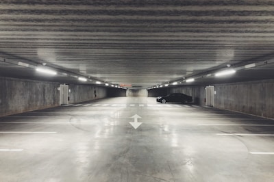 A large, empty underground parking garage with concrete walls and ceiling. There are fluorescent lights lining the ceiling, casting a dull glow throughout the space. A single black car is parked on the right side, while the rest of the parking spots are vacant. The floor is smooth and polished, with directional arrows painted on it, contributing to an overall sterile and industrial atmosphere.