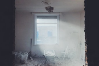 A dimly lit, partially demolished room with a window allowing in some light. Debris is scattered across the floor, and a wheelbarrow filled with rubble sits in the center. On the left, bags filled with debris are visible, while part of the ceiling is damaged, with insulation exposed. The walls are partially unfinished, and some tools are leaning against the right wall.