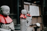 Three stone statues of Buddhist figures wearing red cloths around their necks, arranged in a row. The background features a wooden structure with Japanese writing on a plaque. A white flower is visible near the base of one of the statues.