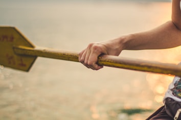 A close-up of a person's hand grasping a wooden paddle with a yellow blade. The background is a softly focused body of water reflecting the golden hues of sunset.