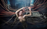 A person wrapped in rope is surrounded by strands of rope extending outward in various directions. The setting appears mysterious and shadowed, with industrial machinery in the background. The figure has striking makeup, with vibrant eye colors, adding to the surreal and artistic atmosphere.