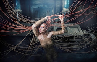 A person wrapped in rope is surrounded by strands of rope extending outward in various directions. The setting appears mysterious and shadowed, with industrial machinery in the background. The figure has striking makeup, with vibrant eye colors, adding to the surreal and artistic atmosphere.