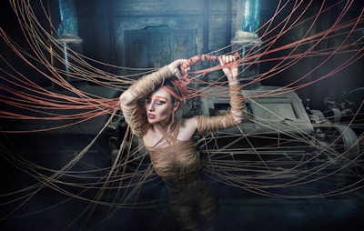 A person wrapped in rope is surrounded by strands of rope extending outward in various directions. The setting appears mysterious and shadowed, with industrial machinery in the background. The figure has striking makeup, with vibrant eye colors, adding to the surreal and artistic atmosphere.