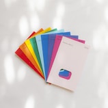 A collection of colorful paint swatches is fanned out on a white surface. Each swatch displays a distinct color and is labeled with a color name and code. The swatches include bright shades of red, orange, yellow, green, blue, purple, pink, and a neutral shade. The lighting creates soft shadows and highlights on the surface.