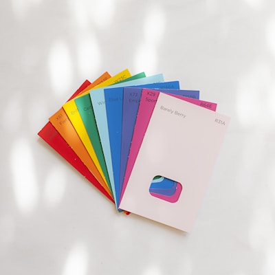 A collection of colorful paint swatches is fanned out on a white surface. Each swatch displays a distinct color and is labeled with a color name and code. The swatches include bright shades of red, orange, yellow, green, blue, purple, pink, and a neutral shade. The lighting creates soft shadows and highlights on the surface.