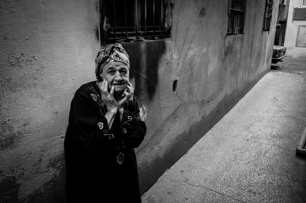 grayscale photography of woman with both hands on cheek standing beside wall