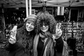 Two people smile and pose with peace signs in front of booths displaying an array of jewelry, likely at a market or fair. One wears a knit hat and has long hair, while the other sports curly hair, sunglasses, and a scarf.