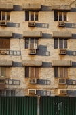 A building facade with multiple windows, each equipped with an air conditioning unit. The wall is made of beige bricks, and there are shadows cast on the surface, indicating a low angle of sunlight. The lower part of the image shows a green corrugated metal fence.