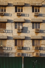 A building facade with multiple windows, each equipped with an air conditioning unit. The wall is made of beige bricks, and there are shadows cast on the surface, indicating a low angle of sunlight. The lower part of the image shows a green corrugated metal fence.