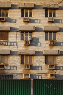 A building facade with multiple windows, each equipped with an air conditioning unit. The wall is made of beige bricks, and there are shadows cast on the surface, indicating a low angle of sunlight. The lower part of the image shows a green corrugated metal fence.