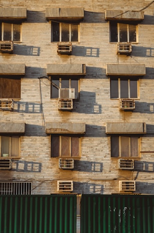 A building facade with multiple windows, each equipped with an air conditioning unit. The wall is made of beige bricks, and there are shadows cast on the surface, indicating a low angle of sunlight. The lower part of the image shows a green corrugated metal fence.