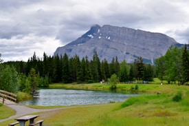 A picturesque landscape featuring a calm lake surrounded by lush green grass and dense evergreen forests. In the background, a majestic mountain with patches of snow stands under a cloudy sky. A wooden bridge crosses a small section of the lake, and a picnic table is positioned in the foreground.