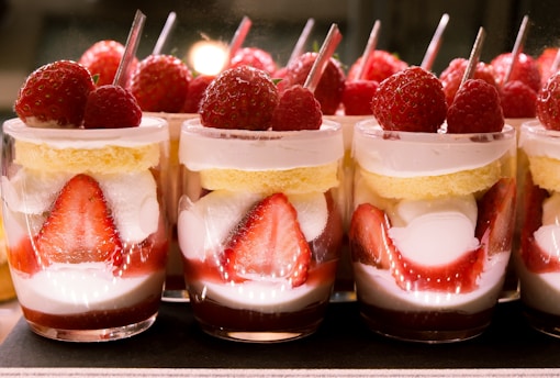Individual dessert cups filled with layers of sliced strawberries, cream, and sponge cake. Each cup is elegantly topped with whole strawberries and raspberries, accompanied by a small spoon.