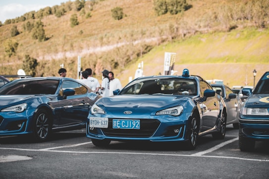 A car meet scene with several blue sports cars parked in a lot. People are gathered around, engaging in conversation under a clear sky and a backdrop of grassy hills.